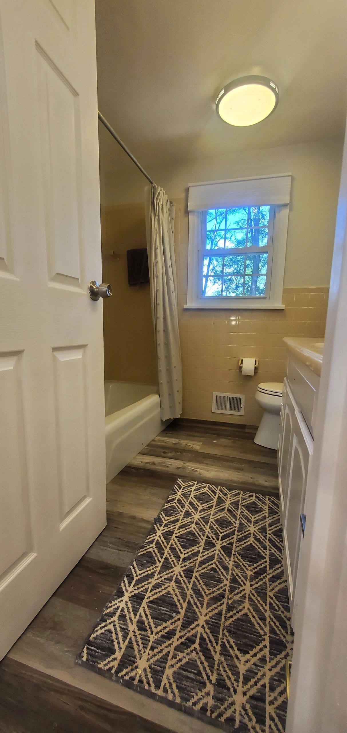 A small bathroom with a patterned rug and a window showing trees outside.