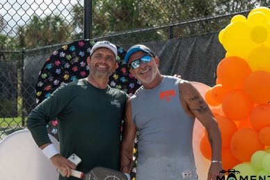 Two men smiling and posing near colorful balloons and a tennis racket.