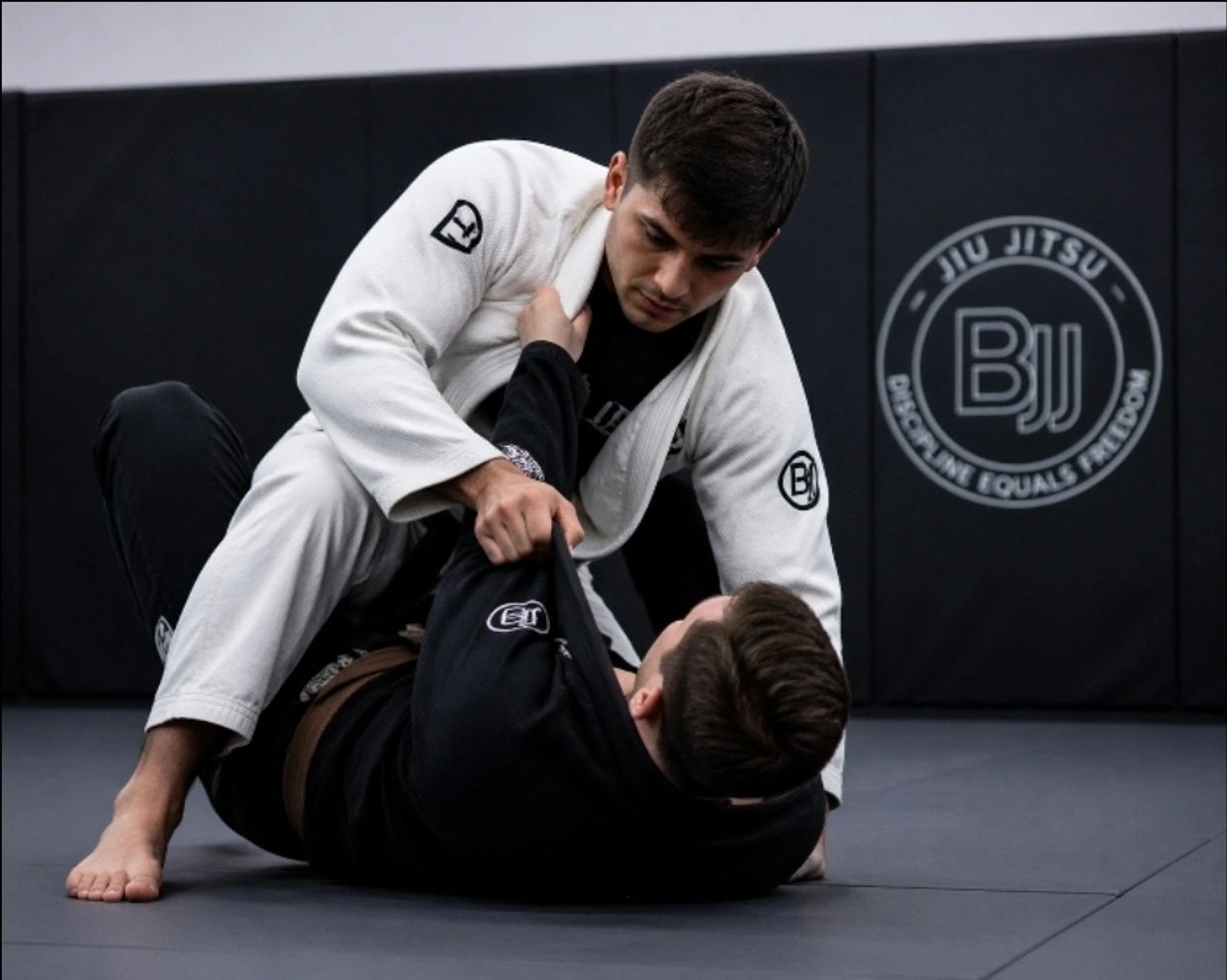 Two men practicing Brazilian Jiu-Jitsu in a gym with BJJ logo.