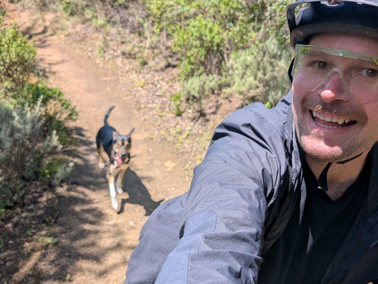 Man wearing helmet and glasses takes selfie with dog on sunny trail.