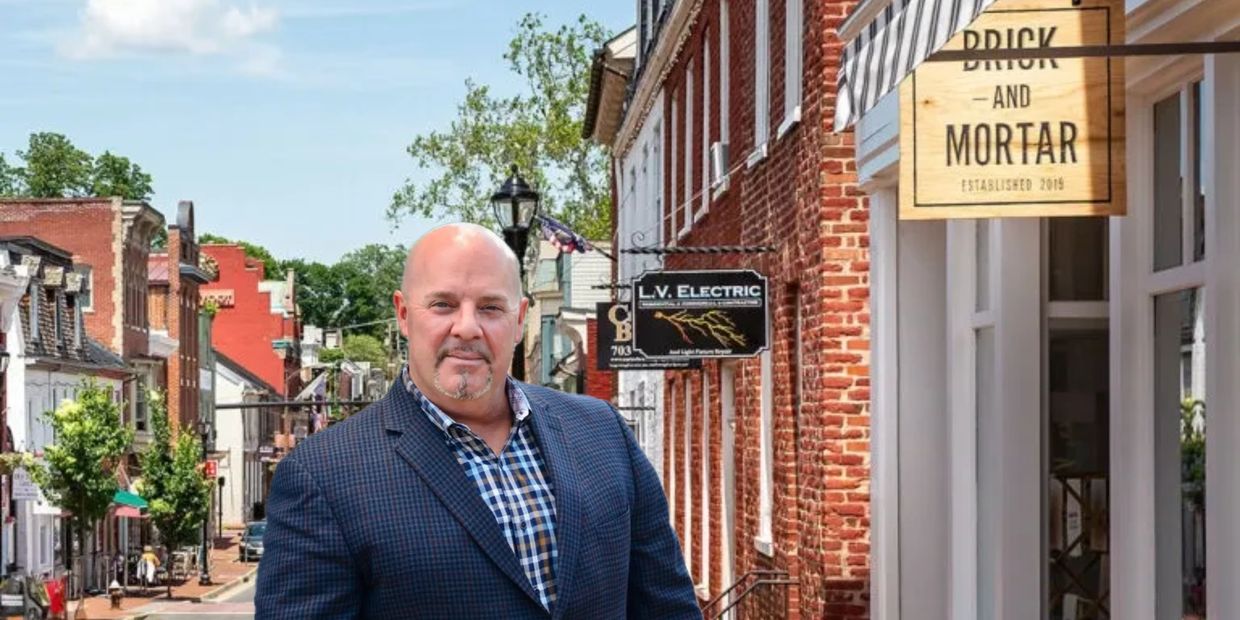 Man in a checkered shirt and blazer stands on a quaint brick-lined street.