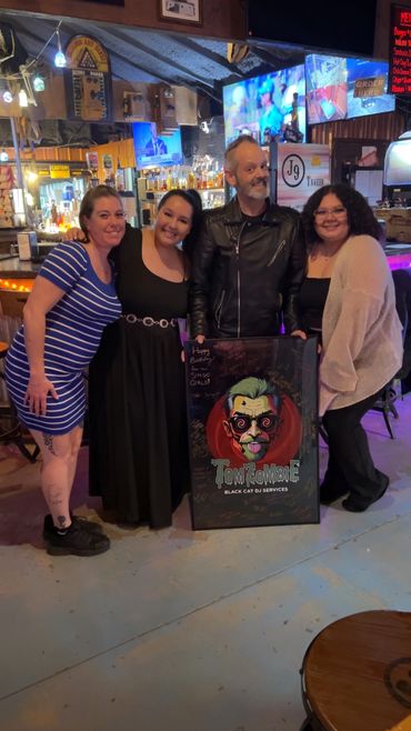 Four people posing happily in a bar with a colorful Tom Zombie poster.