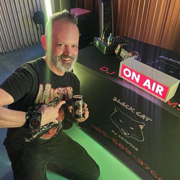Man smiling and holding a BrewDog can near an 'ON AIR' sign in a DJ setup area.