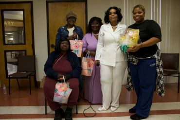 Group of five women holding gift bags and a wrapped bouquet indoors.
