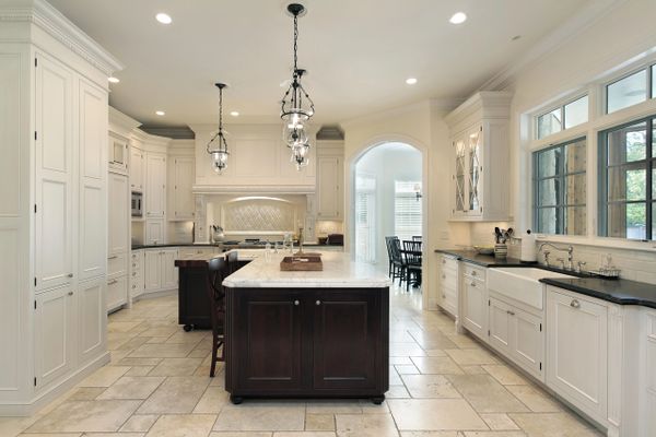 All white kitchen with a dark brown island, light brown tile floor and chandeliers.