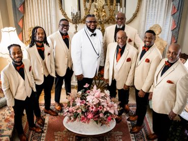 Group of men in beige suits and one in a white suit posing indoors.