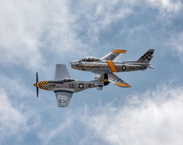 Two vintage military planes flying side by side against a cloudy sky.