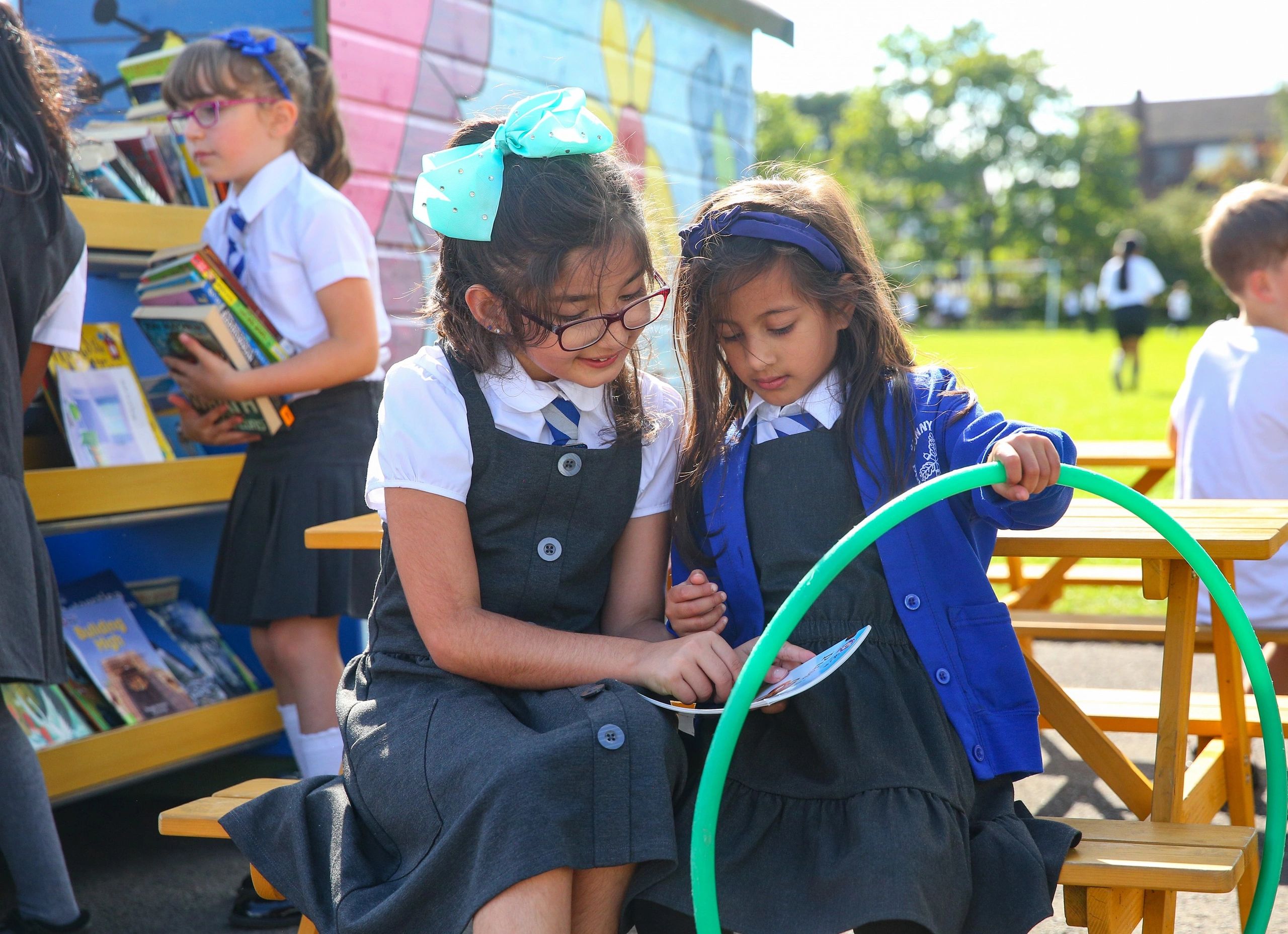 Two schoolgirls in uniform reading together outdoors with a colorful bookshelf behind them.