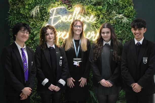A group of students and a teacher posing in front of a neon sign with greenery.