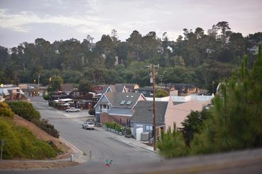 Small town street with houses and greenery in the background under a cloudy sky.