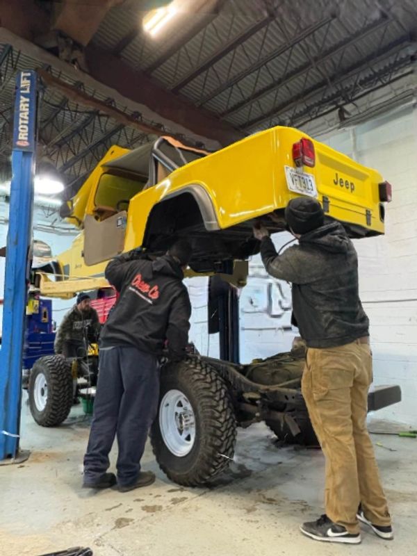 Two men work on reassembling a yellow Jeep body onto its chassis in a garage.