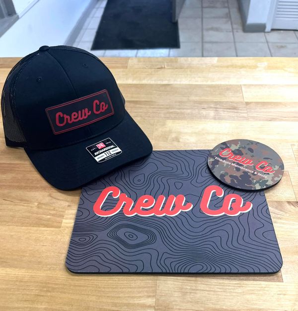 Crew Co branded black cap, mousepad, and camo coaster on a wooden table.