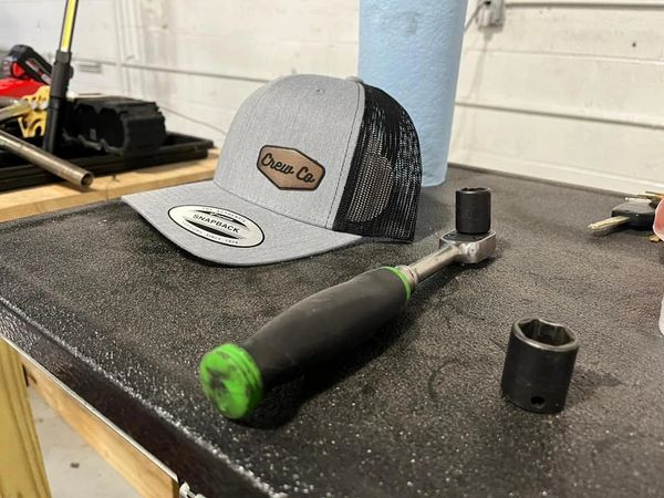 A gray Crew Co snapback hat and a ratchet wrench on a black workbench.