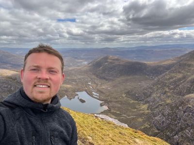 Man taking a selfie on a mountain with a lake and hills in the background.