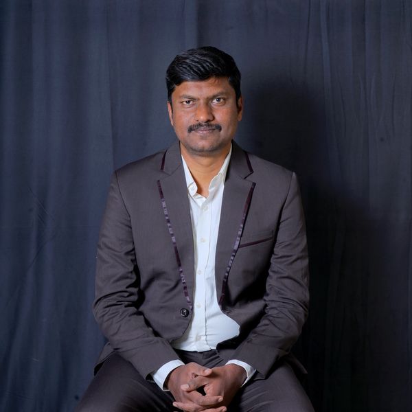 Man in a brown suit sitting against a dark backdrop.
