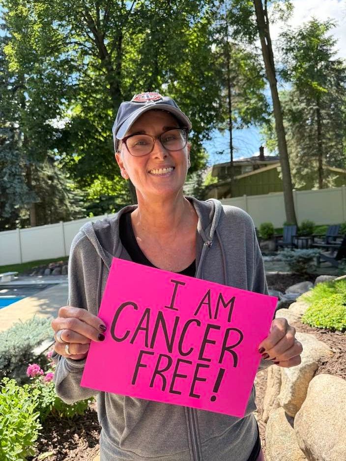 Woman smiling outdoors holding a sign saying 'I AM CANCER FREE!'