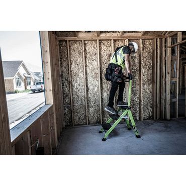 Worker using a green step ladder inside a wooden frame house under construction.