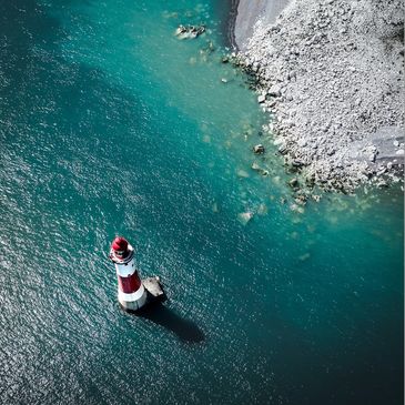 Aerial view of a red and white lighthouse in turquoise water near a rocky shore.
