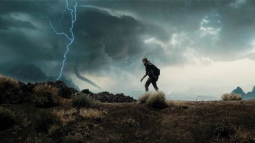 A person walks near a storm with lightning and a tornado in the background.