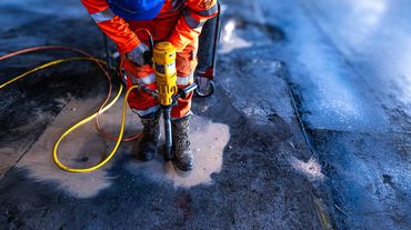 Worker using a jackhammer on a wet concrete floor wearing orange safety gear.