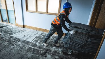 Worker in safety gear pushing a cart stacked with tiles inside a building.