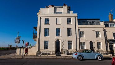 A white multi-story building with a blue convertible car parked outside on a sunny day.