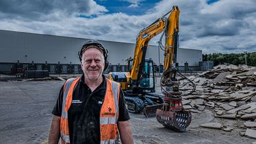 Smiling construction worker in safety gear stands near excavator and debris.