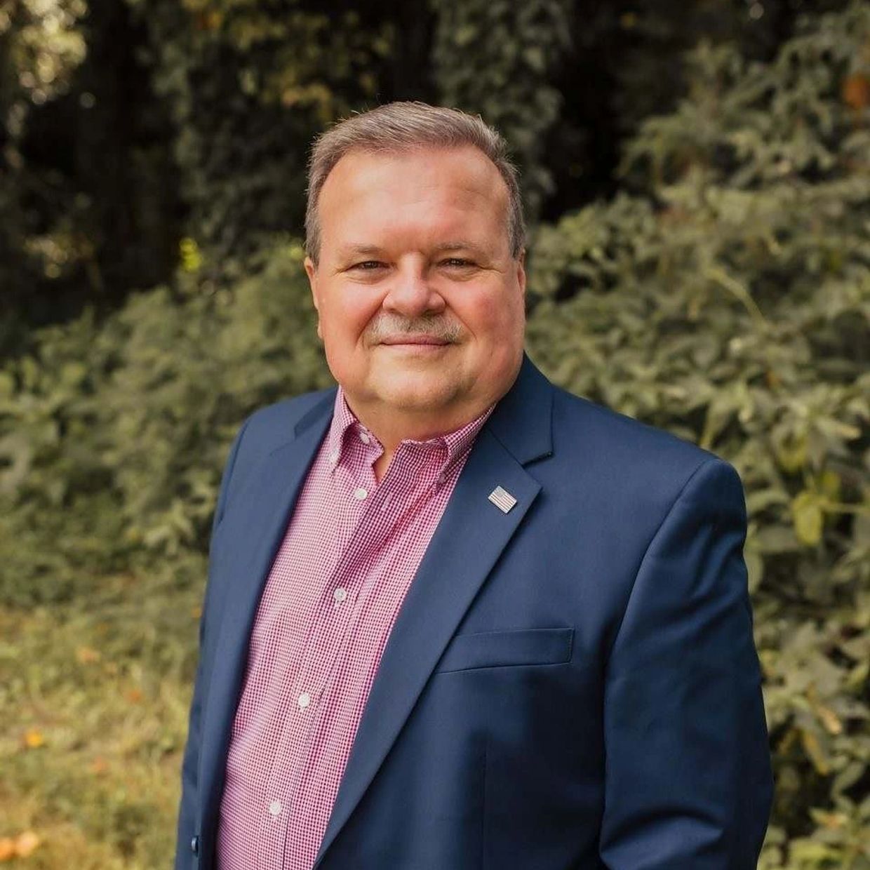 Man in a blue blazer and red checkered shirt smiling outdoors.