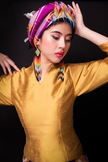 A woman in a golden outfit with colorful beaded earrings and a purple head wrap poses gracefully.