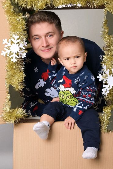 Father and child in festive sweaters posing through decorated cardboard frame.