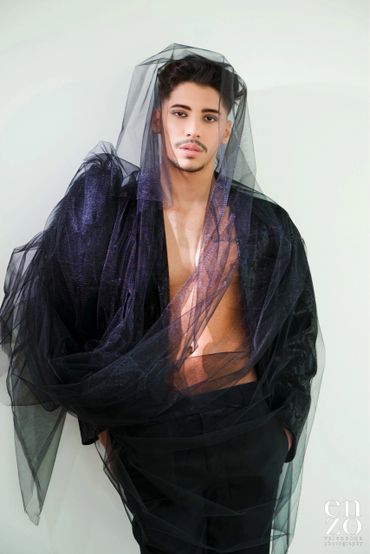 Man draped in sheer black fabric with intense gaze and stylish hair.
