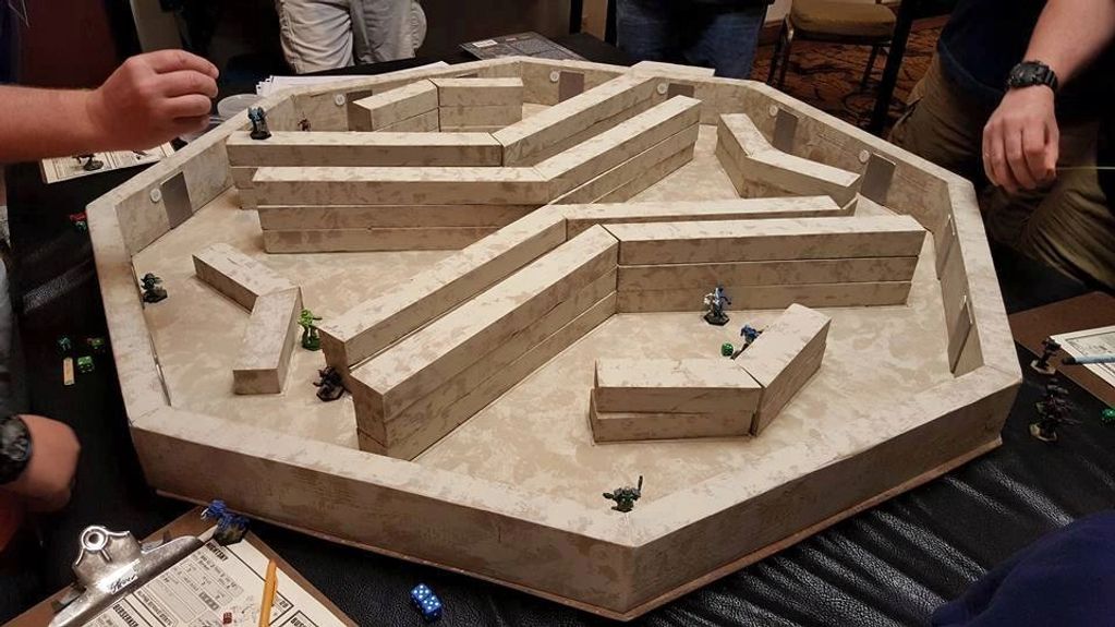 Hands moving pieces on a tabletop game with a maze-like board and miniatures.