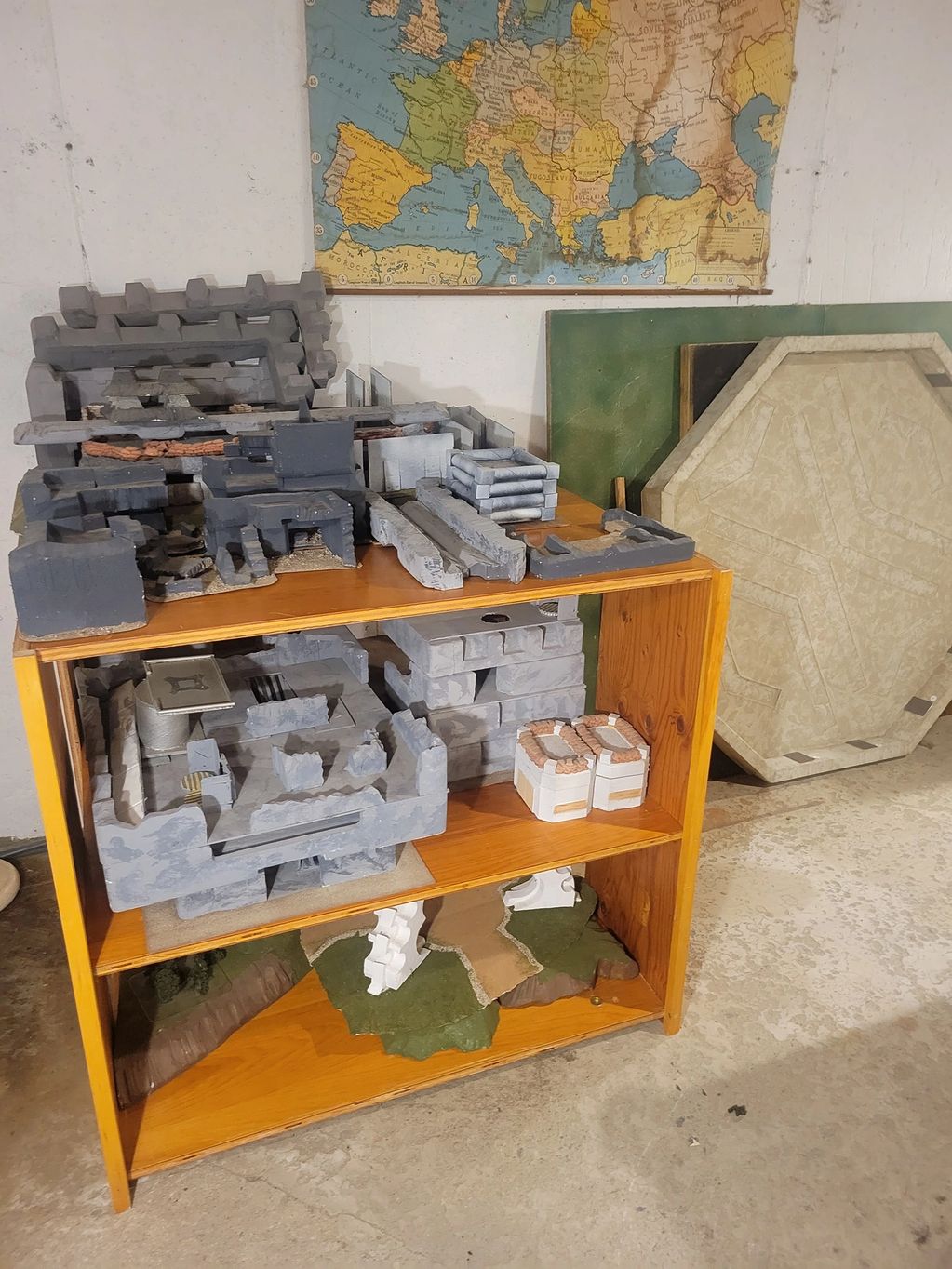 Miniature terrain pieces for tabletop gaming on wooden shelves beneath a map of Europe.