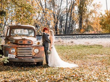 Bride and groom kissing beside a vintage truck in autumn scenery.