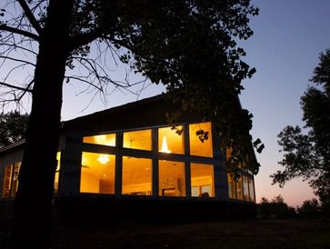 Modern building with large windows lit from inside at twilight, silhouettes of trees around.