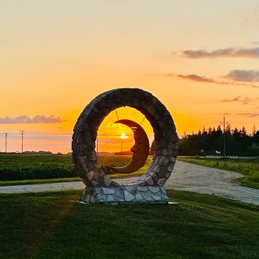 Sunset through a circular stone sculpture with a moon carving in the center of it, on a rural road.