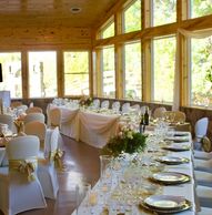 Elegant dining setup in a wooden banquet hall with large windows, decorated tables, and white chairs.