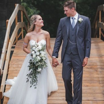 A bride in a white gown and a groom in a gray suit holding hands on a wooden bridge.