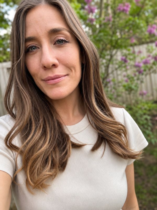 Close-up selfie of a woman with long hair outdoors with flowers in the background.