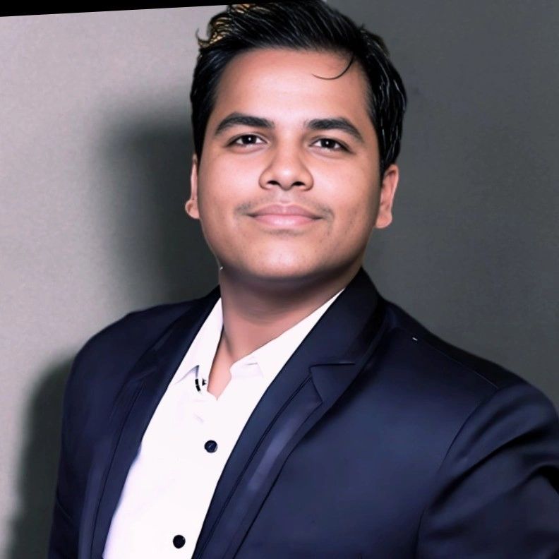 A confident young man in a dark suit and white shirt smiles gently.