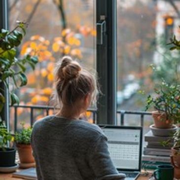 Person working on laptop by a window surrounded by plants.