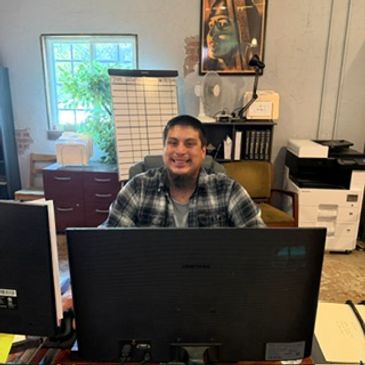 A man smiling behind a large computer monitor at a desk.