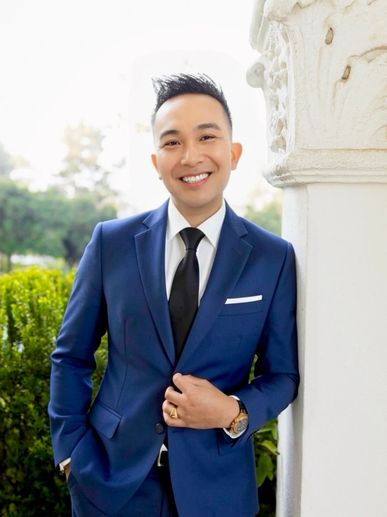 Smiling man in a blue suit leaning against a white column.