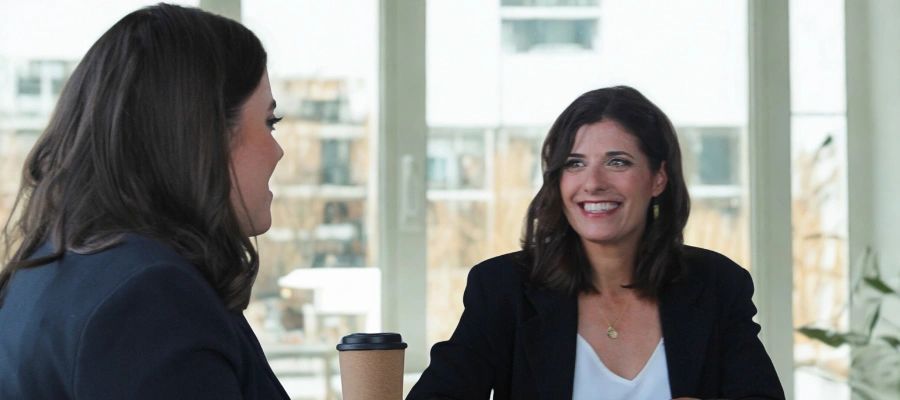 two women sitting at a table with large windows behind them.
