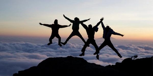 Four people jump joyfully on a mountain against a sunset sky above the clouds.