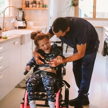 A man affectionately playing with a laughing girl in a wheelchair in a bright kitchen.