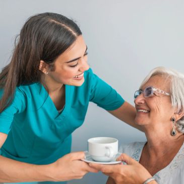 A young nurse smiling while handing a cup of tea to an elderly woman.