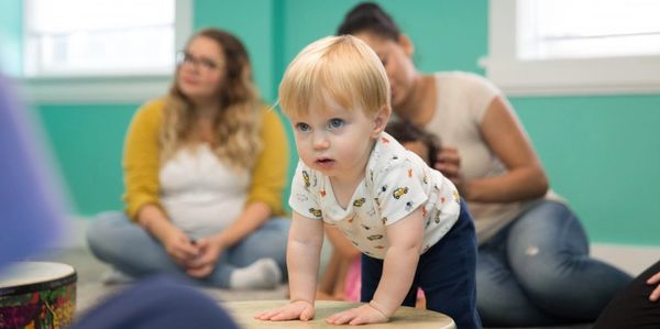 A toddler leaning on a drum with two adults sitting in the background.