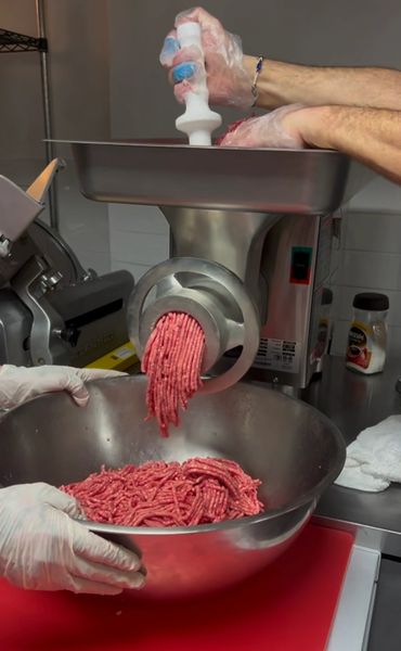 Hands using a meat grinder to mince meat into a bowl in a kitchen.