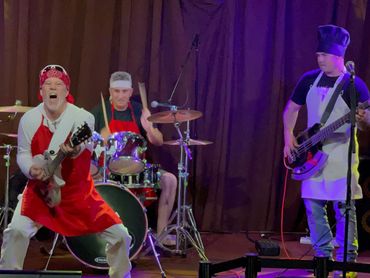 Band members dressed as chefs play guitar, drums, and bass on stage.
